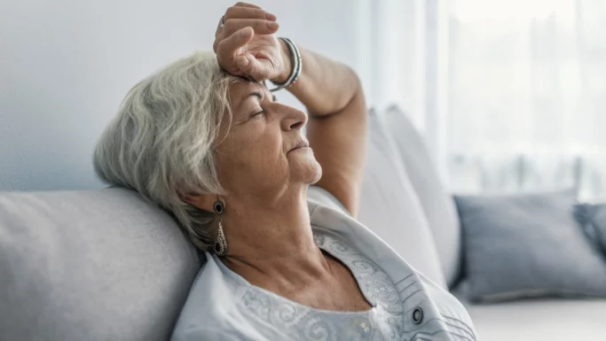 En ældre kvinde med gråt hår sidder i en sofa med lukkede øjne og tilbagelænet hoved, mens hun hviler sig og forsøger at få kræfterne tilbage i det svagt oplyste rum.