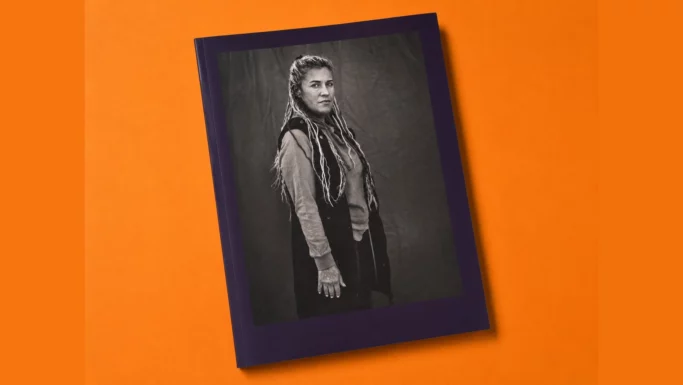 En fotobog med et mørkt omslag, der viser et sort-hvidt portræt af en kvinde med langt hår, der står og ser ind i kameraet, ligger på en orange overflade.