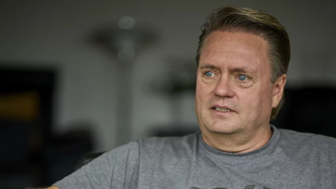 En mand i midten af livet med lyst hår, iført en grå T-shirt, sidder indenfor. Han har blå øjne og et alvorligt udtryk i ansigtet og kigger let til siden, hvilket minder om Kandis-Johnny. Baggrunden er uskarp.