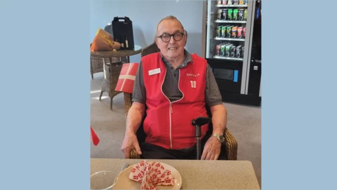 En ældre mand ved navn Keld, iført en rød vest, sidder ved et bord med et dansk flag og en fødselsdagskage – måske for at fejre, at han er gået på pension. I baggrunden står der drikkevarer i en automat, hvilket bidrager til den festlige stemning efter mange års arbejde.