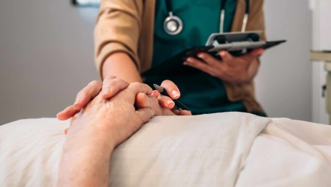 En sundhedsmedarbejder med et stetoskop trøster en patient ved at holde vedkommendes hånd, mens vedkommende tager noter på et skriveunderlag. Fokus er på hænderne, hvilket signalerer empati og omsorg i en medicinsk sammenhæng – en scene, der afspejler den medfølende støtte, man ser i debatten om aktiv dødshjælp i Frankrig.