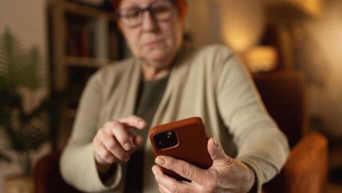 En ældre kvinde med briller sidder indenfor og kigger på sin smartphone, mens hun trykker på skærmen med en finger – måske læser hun eller sletter beskeder.