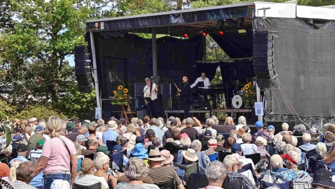 Et band optræder på en udendørs scene, der er pyntet med solsikker, ved Seniorfestivalen, mens et stort publikum sidder og ser på under solrige himmel. Området er omgivet af træer, og mange tilskuere i hatte og sommertøj tager billeder af den livlige scene.