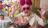 En ældre kvinde med lyserøde, hjerteformede briller og en stor lyserød sløjfe smiler med hænderne løftet bag en kage med 100 stearinlys, mens hun fejrer sin 100-års fødselsdag – en glædelig påmindelse om at tjekke alderen og reflektere over den forventede levealder.