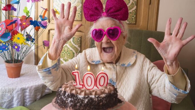 En ældre kvinde med lyserøde, hjerteformede briller og en stor lyserød sløjfe smiler med hænderne løftet bag en kage med 100 stearinlys, mens hun fejrer sin 100-års fødselsdag – en glædelig påmindelse om at tjekke alderen og reflektere over den forventede levealder.
