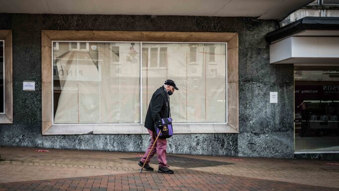 En ældre mand iført en kasket og mundbind går med en stok på en fortovsflise foran tomme butiksvinduer, hvilket afspejler den butiksdød, som mange mindre byer står over for. Han bærer en lilla taske og er klædt i mørkt tøj med lyserøde bukser.