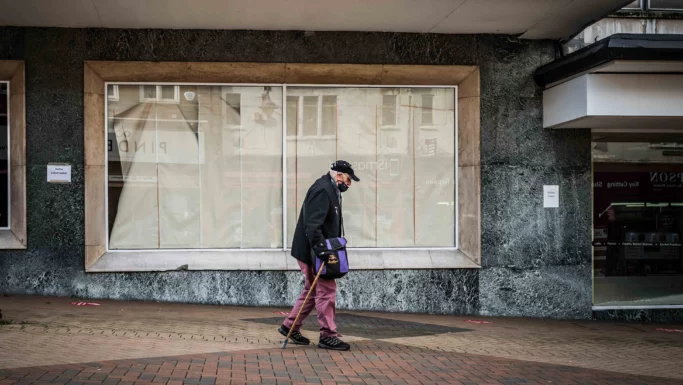 En ældre mand iført en kasket og mundbind går med en stok på en fortovsflise foran tomme butiksvinduer, hvilket afspejler den butiksdød, som mange mindre byer står over for. Han bærer en lilla taske og er klædt i mørkt tøj med lyserøde bukser.