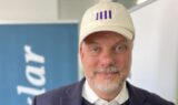 En mand med et lyst skæg og overskæg har en hvid kasket med et blåt logo, en marineblå blazer og en hvid skjorte på. Han står indendørs foran blå og hvide bannere på Seniorfestivalen, hvor der foregår diskussioner om, hvordan man kan arbejde længere eller gå ned i tid.