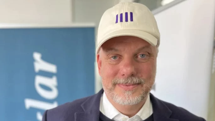 En mand med et lyst skæg og overskæg har en hvid kasket med et blåt logo, en marineblå blazer og en hvid skjorte på. Han står indendørs foran blå og hvide bannere på Seniorfestivalen, hvor der foregår diskussioner om, hvordan man kan arbejde længere eller gå ned i tid.