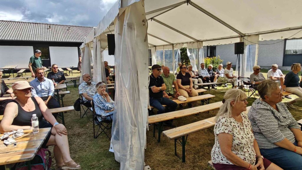 Under Seniorfestivalen sidder folk på bænke under et stort hvidt telt og nyder arrangementet i højt humør. Nogle snakker, mens andre kigger fremad, og et par stykker tager billeder for at huske denne solrige og hyggelige dag.