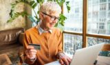 En ældre kvinde med kort, gråt hår og briller smiler, mens hun bruger en bærbar computer til at betale for varer uden for EU og holder et kreditkort i den ene hånd. Hun sidder i et lyst, moderne rum med store vinduer og grønne planter.