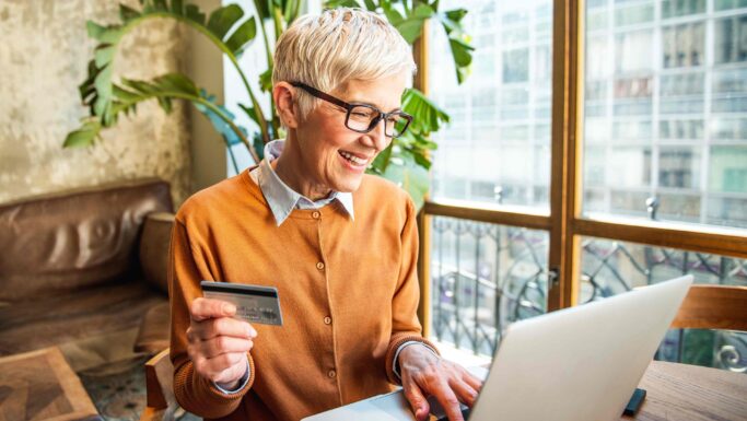 En ældre kvinde med kort, gråt hår og briller smiler, mens hun bruger en bærbar computer til at betale for varer uden for EU og holder et kreditkort i den ene hånd. Hun sidder i et lyst, moderne rum med store vinduer og grønne planter.