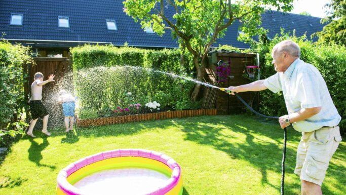 En ældre mand sprøjter legende vand på to børn med en haveslange ved siden af en lille oppustelig pool i en solrig baghave, hvor han nyder sommeren i fulde drag. Børnene griner og dækker sig for vandet, mens de køler af under en af sæsonens hedebølger. Træer og farvestrålende blomster omgiver den muntre scene.