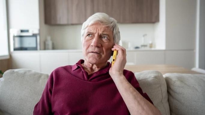 En ældre mand med gråt hår, iført en rødbrun skjorte, sidder i en sofa og taler i en gul smartphone, mens han ser bekymret ud – måske diskuterer han et problem med sit elselskab eller håber på at finde billigere strøm. I baggrunden ses et moderne køkken.