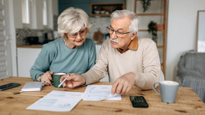 Et ældre ægtepar sidder ved et bord og gennemgår papirer sammen, muligvis for at drøfte pensionsspørgsmål. Manden peger på et dokument med en pen, mens kvinden holder en kop. Der ligger også en lommeregner og en notesblok på bordet, hvilket tyder på, at de måske er i gang med at beregne erstatning fra pensionsselskabet eller planlægger deres økonomi med henblik på pensionering.