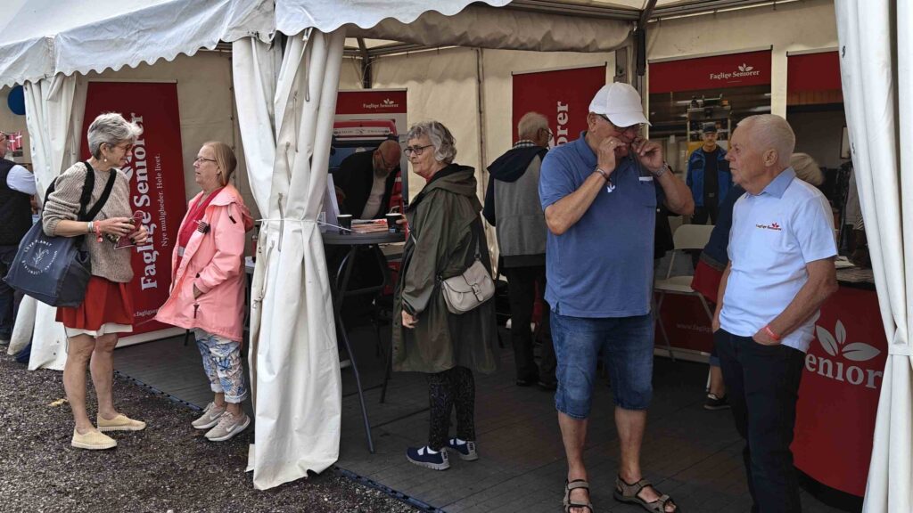 En gruppe ældre står og snakker under et hvidt telt på Seniorfestivalen, alle i højt humør, med informationsborde og bannere i baggrunden.