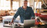 En pensionist i en denimskjorte står i et værksted og kigger på en bærbar computer, der ligger på en arbejdsbænk. I nærheden ligger værktøj, en kop og et par briller, mens der i baggrunden ses træbearbejdningsudstyr, og han overvejer, om han skal arbejde videre.