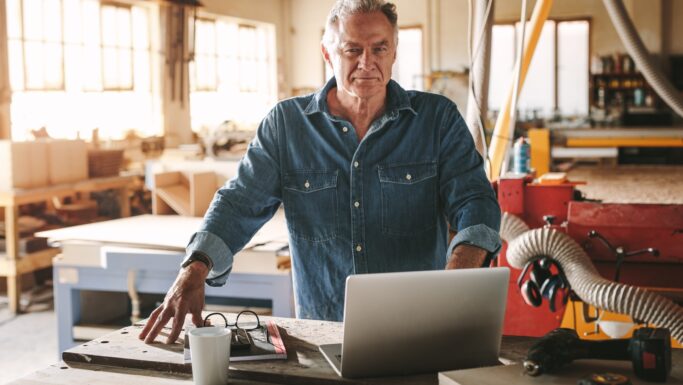 En pensionist i en denimskjorte står i et værksted og kigger på en bærbar computer, der ligger på en arbejdsbænk. I nærheden ligger værktøj, en kop og et par briller, mens der i baggrunden ses træbearbejdningsudstyr, og han overvejer, om han skal arbejde videre.