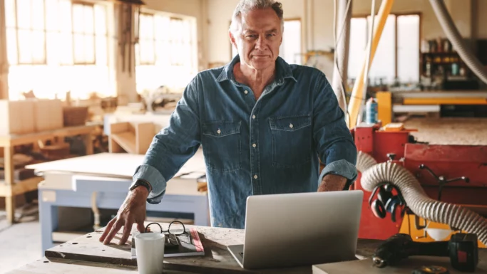 En pensionist i en denimskjorte står i et værksted og kigger på en bærbar computer, der ligger på en arbejdsbænk. I nærheden ligger værktøj, en kop og et par briller, mens der i baggrunden ses træbearbejdningsudstyr, og han overvejer, om han skal arbejde videre.