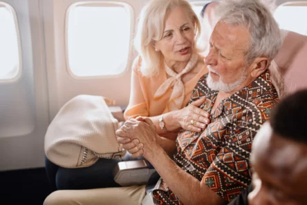 En ældre kvinde trøster en ældre mand, der ser ud til at have det dårligt, mens de sidder sammen på et fly. Kvinden holder ham i hånden og rører ved hans skulder, og man kan tydeligt se bekymringen i hendes ansigt, trods irritationen blandt de andre passagerer.