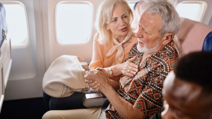 En ældre kvinde trøster en ældre mand, der ser ud til at have det dårligt, mens de sidder sammen på et fly. Kvinden holder ham i hånden og rører ved hans skulder, og man kan tydeligt se bekymringen i hendes ansigt, trods irritationen blandt de andre passagerer.