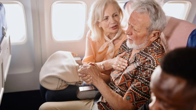 En ældre kvinde trøster en ældre mand, der ser ud til at have det dårligt, mens de sidder sammen på et fly. Kvinden holder ham i hånden og rører ved hans skulder, og man kan tydeligt se bekymringen i hendes ansigt, trods irritationen blandt de andre passagerer.