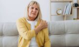 En ældre kvinde med blondt hår, iført en gul cardigan, sidder på en sofa og holder sig smertefuldt om sin venstre skulder. I baggrunden ses en reol med bøger og et ur. Hun ligner en førtidspensionist med smerter.