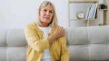 En ældre kvinde med blondt hår, iført en gul cardigan, sidder på en sofa og holder sig smertefuldt om sin venstre skulder. I baggrunden ses en reol med bøger og et ur. Hun ligner en førtidspensionist med smerter.