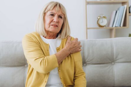 En ældre kvinde med blondt hår, iført en gul cardigan, sidder på en sofa og holder sig smertefuldt om sin venstre skulder. I baggrunden ses en reol med bøger og et ur. Hun ligner en førtidspensionist med smerter.