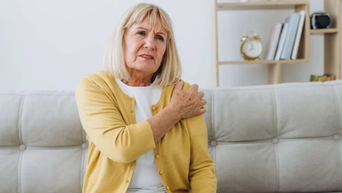 En ældre kvinde med blondt hår, iført en gul cardigan, sidder på en sofa og holder sig smertefuldt om sin venstre skulder. I baggrunden ses en reol med bøger og et ur. Hun ligner en førtidspensionist med smerter.