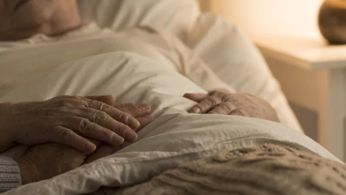 En ældre person ligger i sengen under et cremefarvet tæppe, med hænderne blidt hvilende oven på dynen. Blødt, varmt lys skaber en fredfyldt atmosfære, mens omsorgsfulde hospiceansatte sørger for komfort og værdighed i denne følsomme tid.