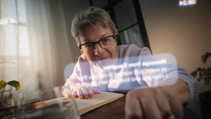 En ældre kvinde med briller sidder ved et bord og rækker sig fremad, mens hun smiler. Hun har en åben bog foran sig, og i forgrunden ses refleksioner af digital tekst og skærmbilledet fra en mobil med en Fup-sms.