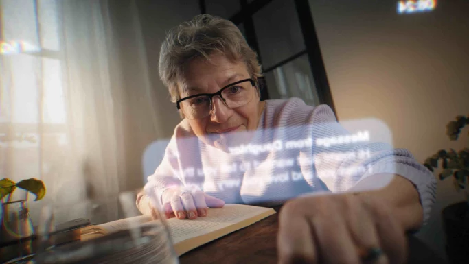 En ældre kvinde med briller sidder ved et bord og rækker sig fremad, mens hun smiler. Hun har en åben bog foran sig, og i forgrunden ses refleksioner af digital tekst og skærmbilledet fra en mobil med en Fup-sms.