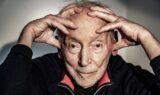 Gregers Dirckinck-Holmfeld, den velkendte tv-vært, sidder med hænderne hvilende på panden. Han har en mørk trøje med rød krave på og ser direkte ind i kameraet med et eftertænksomt udtryk.