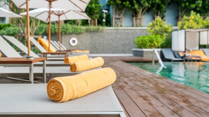 En solsenge ved poolen med et sammenrullet gult håndklæde på, ved siden af en swimmingpool omgivet af flere reserverede liggestole, parasoller og grønne planter på en solrig dag.