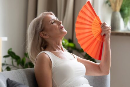 En ældre kvinde i en hvid bluse sidder på en sofa med lukkede øjne og vifter sig med en orange vifte for at beskytte sig mod varmen; hun ser ud til at have det varmt eller være overophedet i et lyst indendørs miljø med planter i baggrunden.