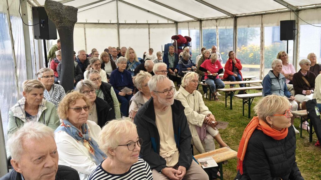 En gruppe ældre sidder på bænke inde i et hvidt telt på Seniorfestivalen og lytter opmærksomt til noget, der ligger uden for synsfeltet. De fleste har briller på og er iført lette jakker, og stemningen er rolig og koncentreret.