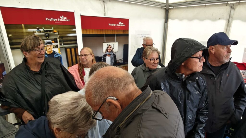 En gruppe ældre, hvoraf mange har regnjakker på, står tæt sammen inde i et telt med bannere fra Faglige Seniorer i baggrunden på Seniorfestivalen. Nogle ser ud til at tale sammen, mens andre ser sig omkring i højt humør.