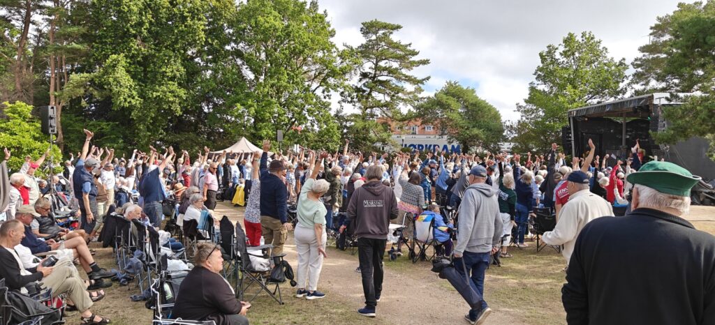 En skare mennesker udendørs, hvoraf mange står med armene løftet mod en scene til højre, udstråler højt humør ved denne livlige seniorfestival. Nogle sidder på klapstole midt i den muntre stemning. Træer omgiver området, og i baggrunden ses et telt. Vejret ser overskyet, men lyst ud – perfekt til at tage billeder af denne glade sammenkomst.