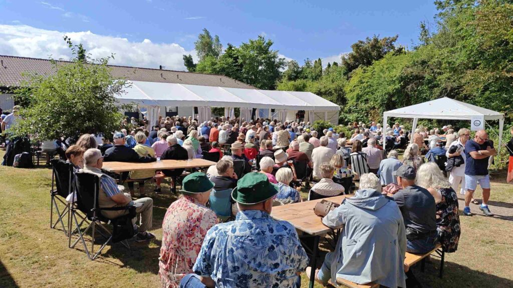 En stor gruppe mennesker sidder ved træborde udendørs med udsigt til et telt, hvor der afholdes et arrangement. Vejret er solrigt, og alle ser ud til at nyde fællesskabet ved Seniorfestivalen i højt humør.