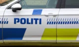 Set fra siden af en hvid politibil med blå og gule markeringer og ordet »POLITI« skrevet på døren, hvilket angiver, at det er et dansk politikøretøj. Billedet ligner en »automatisk kladde«, der gengiver de karakteristiske træk ved danske politibiler.
