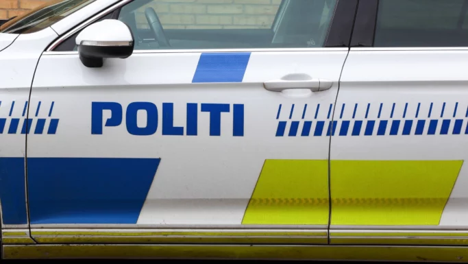 Set fra siden af en hvid politibil med blå og gule markeringer og ordet »POLITI« skrevet på døren, hvilket angiver, at det er et dansk politikøretøj. Billedet ligner en »automatisk kladde«, der gengiver de karakteristiske træk ved danske politibiler.