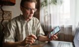 En mand med briller og en ternet skjorte sidder ved et skrivebord med en bærbar computer og holder en smartphone i hånden, hvor der vises et indgående opkald vedrørende MitID. Han ser på telefonen med et koncentreret udtryk, bekymret for, at det måske er svindel, mens han overvejer, om han skal spærre MitID. Sollys strømmer ind gennem et vindue med hvide gardiner.