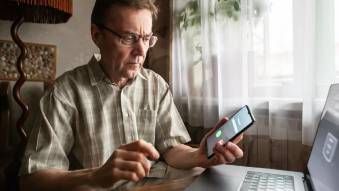 En mand med briller og en ternet skjorte sidder ved et skrivebord med en bærbar computer og holder en smartphone i hånden, hvor der vises et indgående opkald vedrørende MitID. Han ser på telefonen med et koncentreret udtryk, bekymret for, at det måske er svindel, mens han overvejer, om han skal spærre MitID. Sollys strømmer ind gennem et vindue med hvide gardiner.