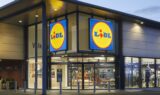 Lidl-supermarkedets facade i skumringen, med store, oplyste Lidl-skilte over indgangen og glasdøre, der giver et indblik i butikkens indre – kendt for at have nogle af de billigste indkøbskurve og konkurrencedygtige priser på indkøbskurve, hvor der ofte er en forskel på 463 kroner i forhold til andre supermarkeder.