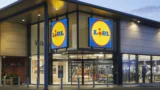 Lidl-supermarkedets facade i skumringen, med store, oplyste Lidl-skilte over indgangen og glasdøre, der giver et indblik i butikkens indre – kendt for at have nogle af de billigste indkøbskurve og konkurrencedygtige priser på indkøbskurve, hvor der ofte er en forskel på 463 kroner i forhold til andre supermarkeder.