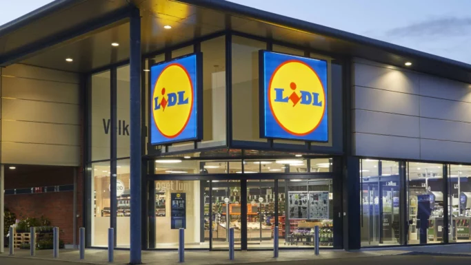 Lidl-supermarkedets facade i skumringen, med store, oplyste Lidl-skilte over indgangen og glasdøre, der giver et indblik i butikkens indre – kendt for at have nogle af de billigste indkøbskurve og konkurrencedygtige priser på indkøbskurve, hvor der ofte er en forskel på 463 kroner i forhold til andre supermarkeder.