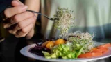 En person, der holder en gaffel med spirer over en tallerken med frisk salat, herunder salatblade, tomat, strimlet rødkål og gul peberfrugt – vær opmærksom på risikoen for salmonella, når du spiser rå spirer.