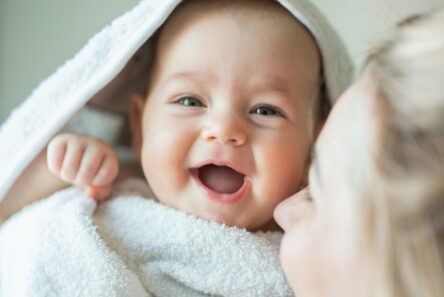 En smilende baby, svøbt i et hvidt håndklæde, kigger ind i kameraet. En voksen med blondt hår, der kun delvist kan ses, står tæt på babyens ansigt. Scenen fremstår varm og munter og indfanger et ømt øjeblik – måske er det de små Svend og Viola, to navne til nyfødte, der vinder indpas i moderne familier.