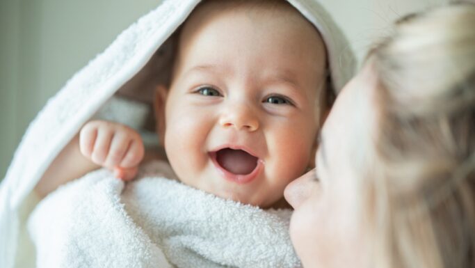 En smilende baby, svøbt i et hvidt håndklæde, kigger ind i kameraet. En voksen med blondt hår, der kun delvist kan ses, står tæt på babyens ansigt. Scenen fremstår varm og munter og indfanger et ømt øjeblik – måske er det de små Svend og Viola, to navne til nyfødte, der vinder indpas i moderne familier.